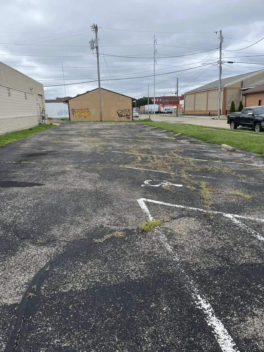 A commercial parking lot before sealcoating, with cracking across the surface, weeds growing through the joints and faded white stall markings